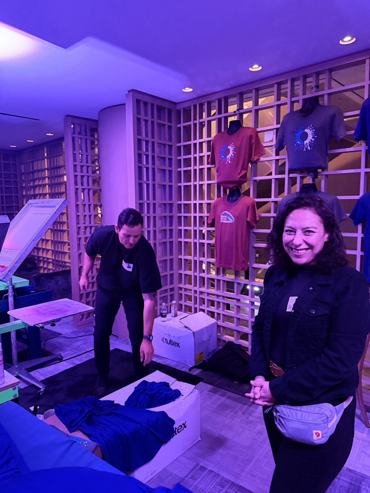 Merch Troop crew member setting up an onsite printing station beneath a lattice display of shirts