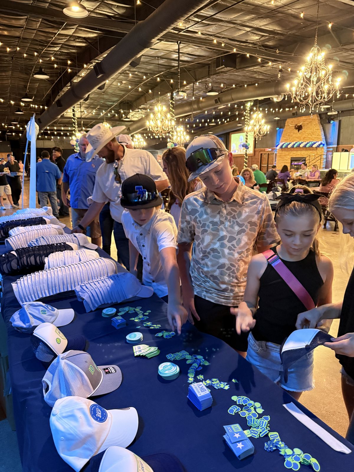 Long table of trucker hats and patch options at an evening event with guests browsing